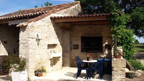 Un edificio de piedra con techo de tejas, un pequeño espacio al aire libre con sillas azules y una mesa, y plantas en macetas.