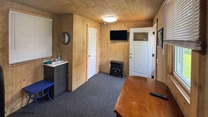 Une pièce aux murs en bois, avec une porte blanche, un téléviseur fixé au-dessus, un banc, une petite table et une fenêtre avec des stores.