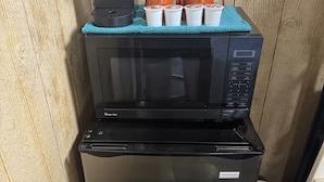 Un petit coin cuisine comprenant un micro-ondes et un réfrigérateur noirs, une machine à café Keurig et quatre gobelets jetables posés sur un tissu de couleur turquoise.