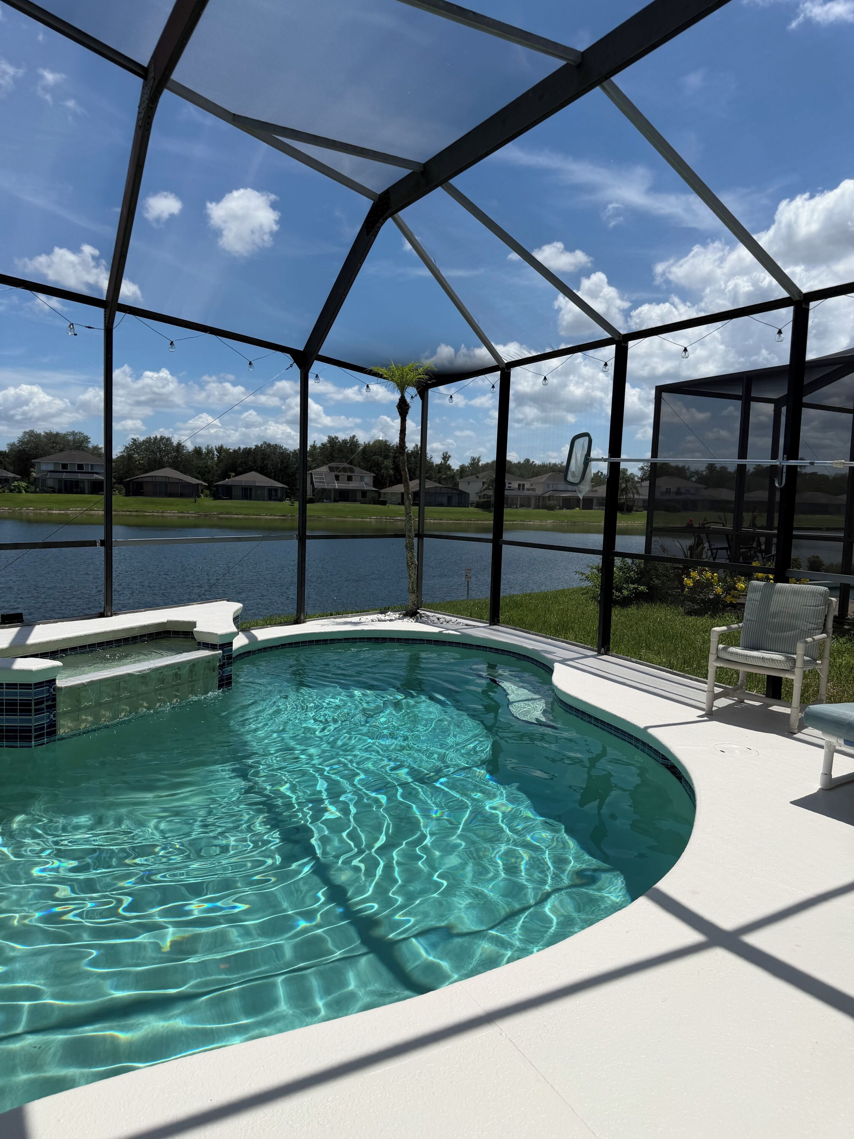 A swimming pool with a clear blue water surface, surrounded by a covered patio area with a view of a lake and houses in the background.
