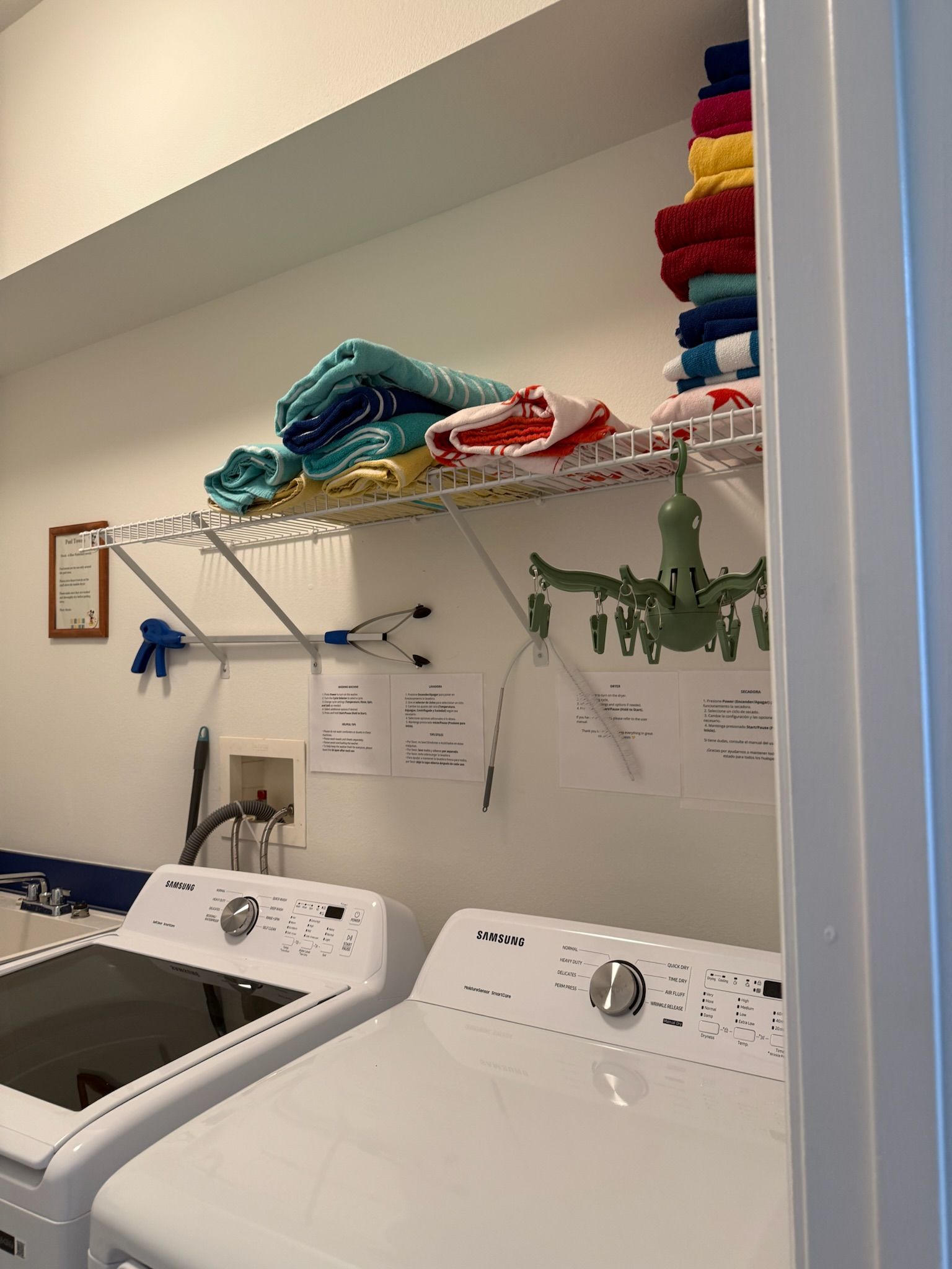 Zona de lavandería con dos lavadoras Samsung, un tendedero con toallas dobladas y un tendedero de pared con perchas.