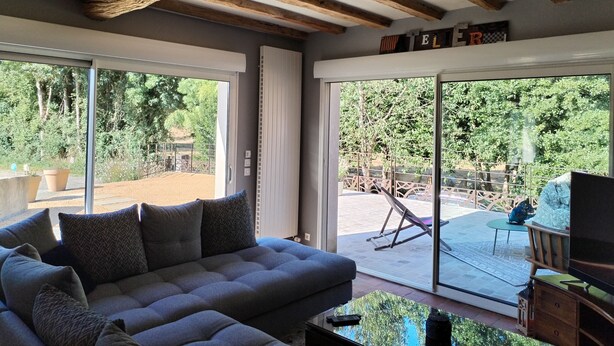 A modern living room with a grey sofa, wooden ceiling beams, large sliding glass doors, and a view of an outdoor area with a swing set. - 8-sleeper country house, 50 minutes from the 24 Hours of Le Mans race track (Saint-Longis)