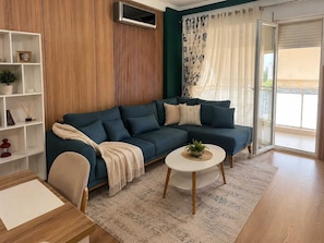 A modern living room with a sectional sofa, a small round coffee table, a white side table, a bookshelf, and a sliding glass door to the outside. - Modern Central Tirana Apartment with Free Parking & Fast WiFi (Tiranë)