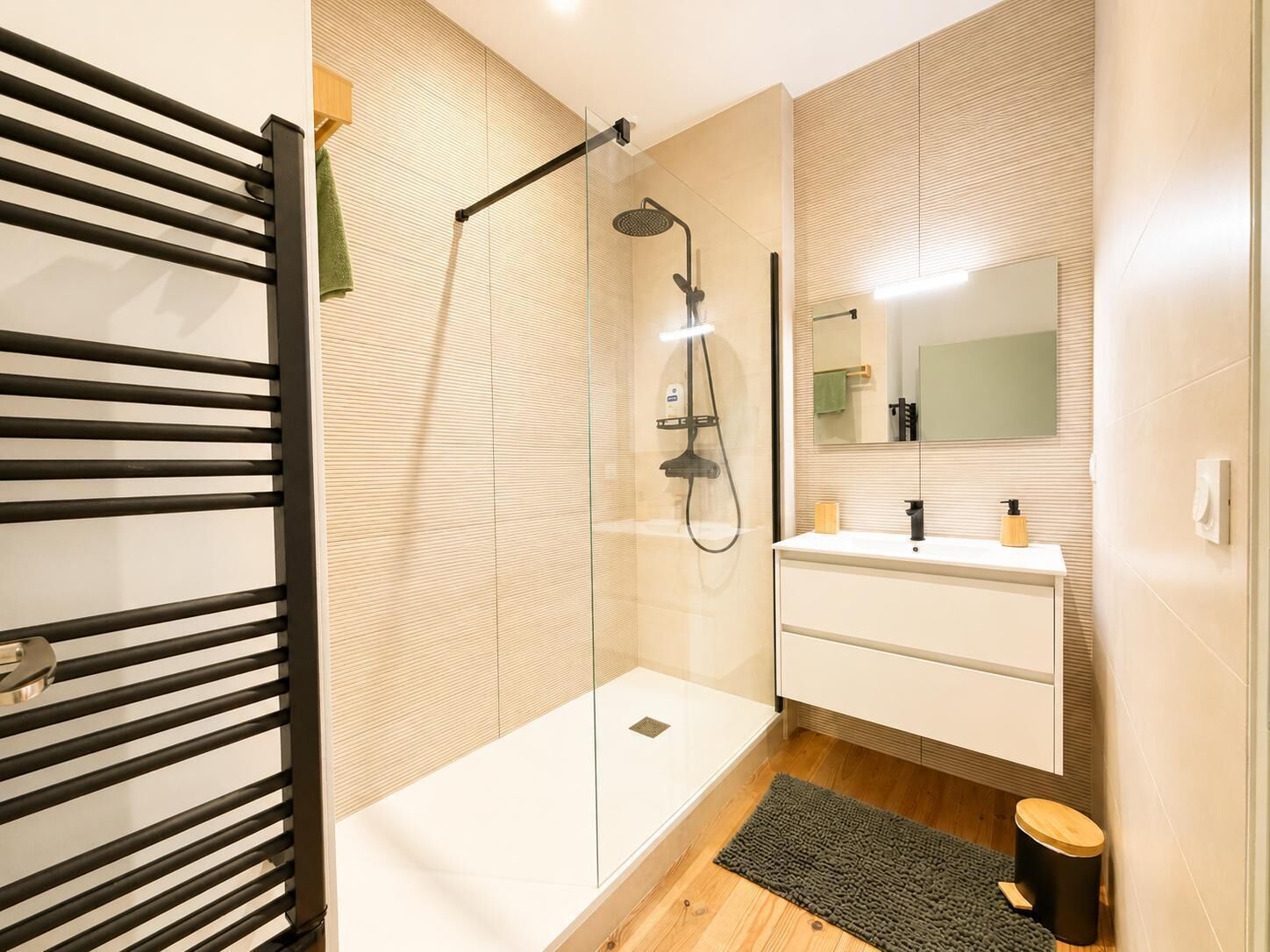 Un baño moderno con cabina de ducha de vidrio, tocador blanco con espejo y suelo de madera.