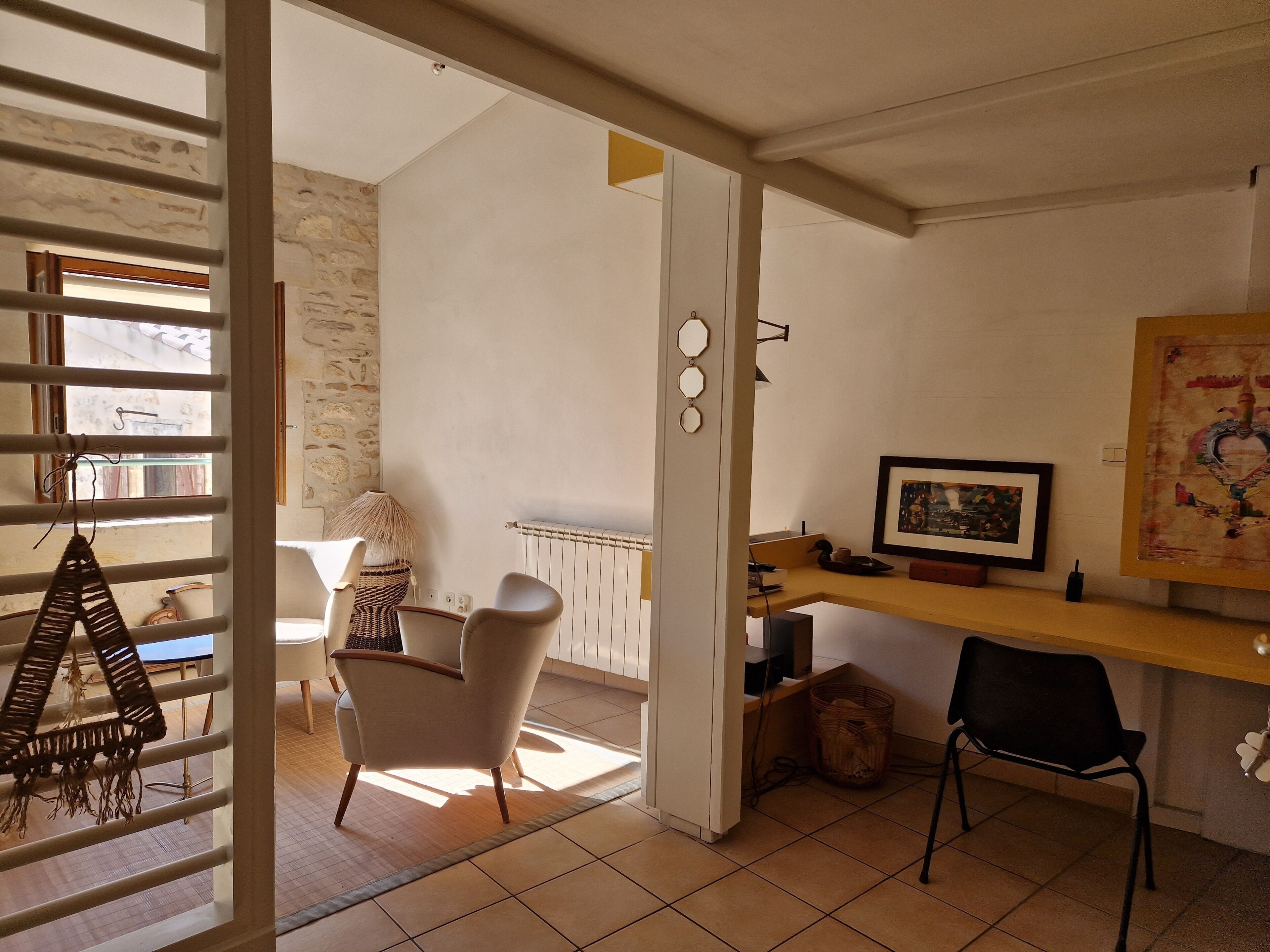 Een kamer met een bureau, stoelen, een radiator, een muur met een schilderij en een raam met jaloezieën.