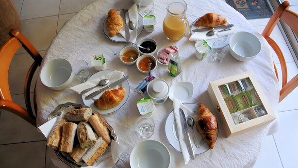 A round dining table with a white tablecloth, surrounded by chairs, and set with breakfast items including croissants, bread, and beverages.