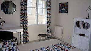 A bedroom with a bed, a desk, a chair, a small table, a radiator, and a bookshelf.