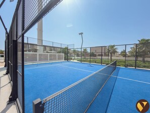 Una cancha de tenis con una red, rodeada por una valla metálica, y un cielo despejado al fondo.