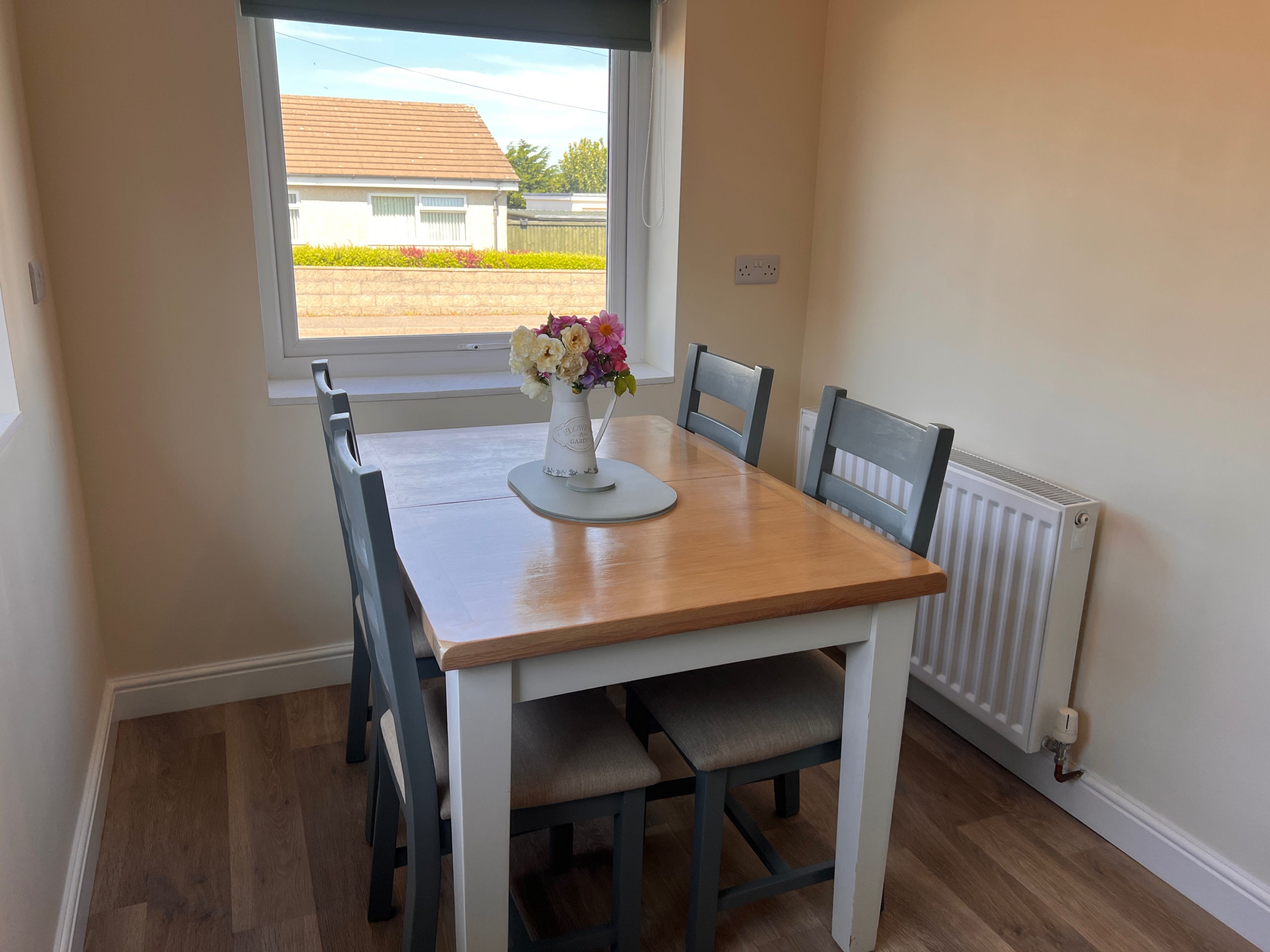 A dining table with chairs, a vase of flowers, and a view of a house through a window.