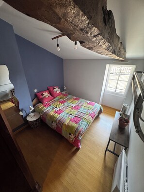 Ein Schlafzimmer mit einem Bett, einem Nachttisch mit Pflanze, einem Fenster mit Gitter und einer Decke mit freiliegendem Holz.