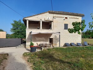Ein zweistöckiges Steinhaus mit überdachter Terrasse, Sitzgelegenheiten im Freien und einem Garten.