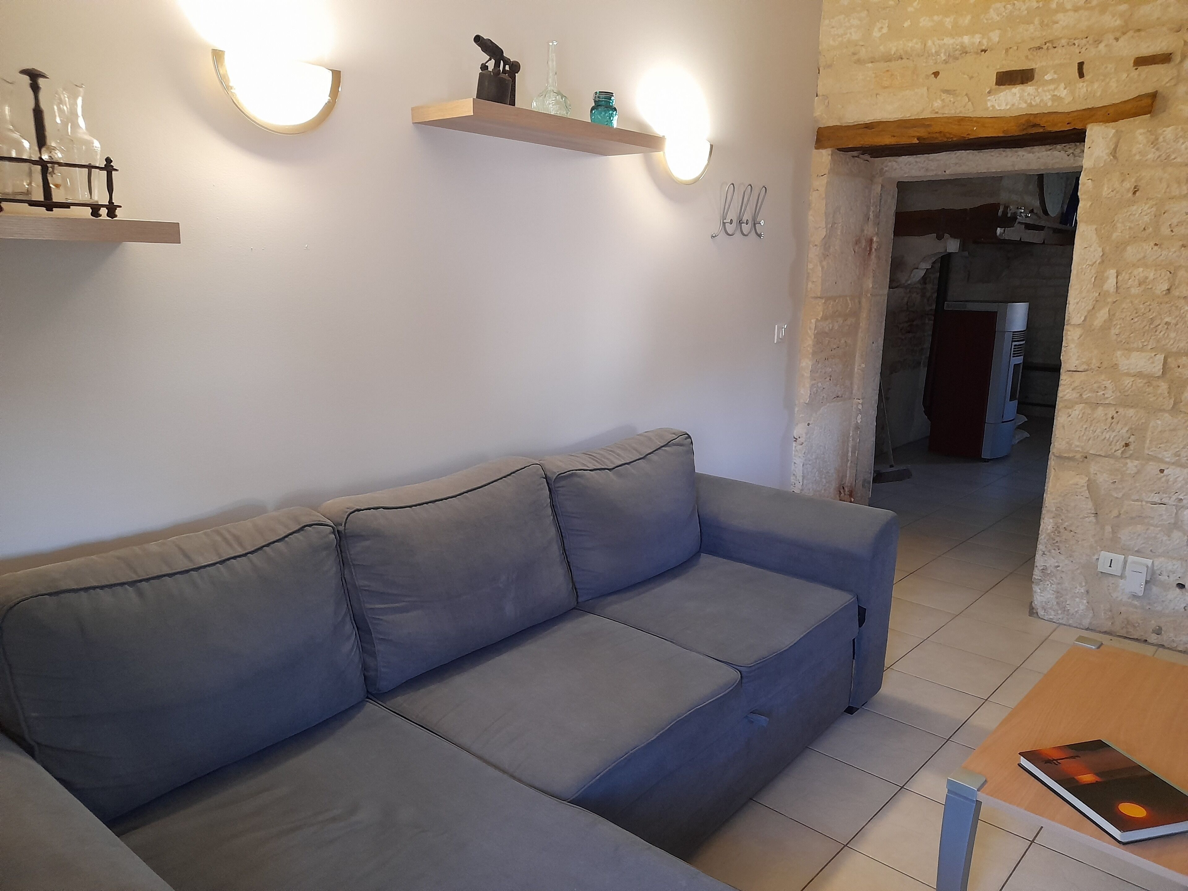 A living room with a grey sofa, wall-mounted shelves with decorative items, and a doorway leading to another room.
