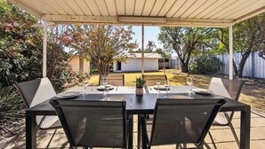 A covered patio with a table set for two, surrounded by chairs, and a view of a backyard with a house and trees. - Modern Family house with BBQ (Dubbo)