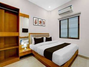 A hotel room with a bed, a wardrobe, a bedside table with a lamp, a wall-mounted TV, framed artwork, an air conditioning unit, and a window with blinds. - Deluxe Hotel O Dashashwamedh Ghat (Varanasi)