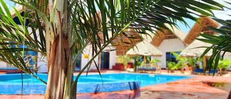 A pool area with palm trees, a swimming pool, and a building with a thatched roof in the background.