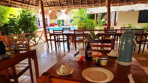 Une table à manger en bois, avec des chaises, une bouteille, un bocal et une assiette, dans un décor tropical avec une piscine et des plantes en arrière-plan.