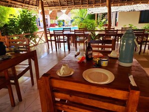 A wooden dining table with chairs, a bottle, a jar, and a plate in a tropical setting with a pool and plants in the background. - Mkeka Spice Lodge Jambian (Unguja South Region)