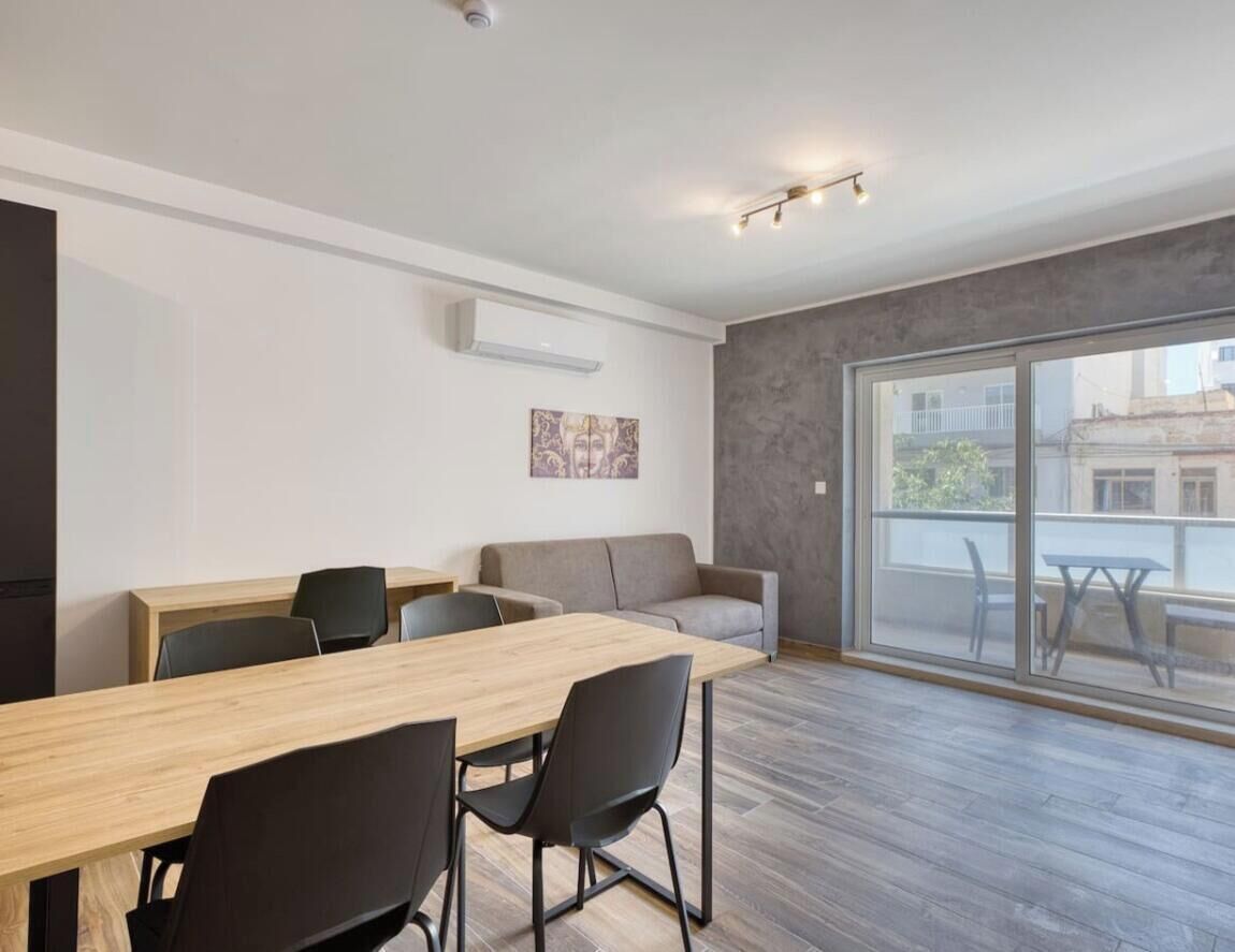 A modern room with a wooden dining table, black chairs, a grey sofa, and a large window offering a view of the outside.