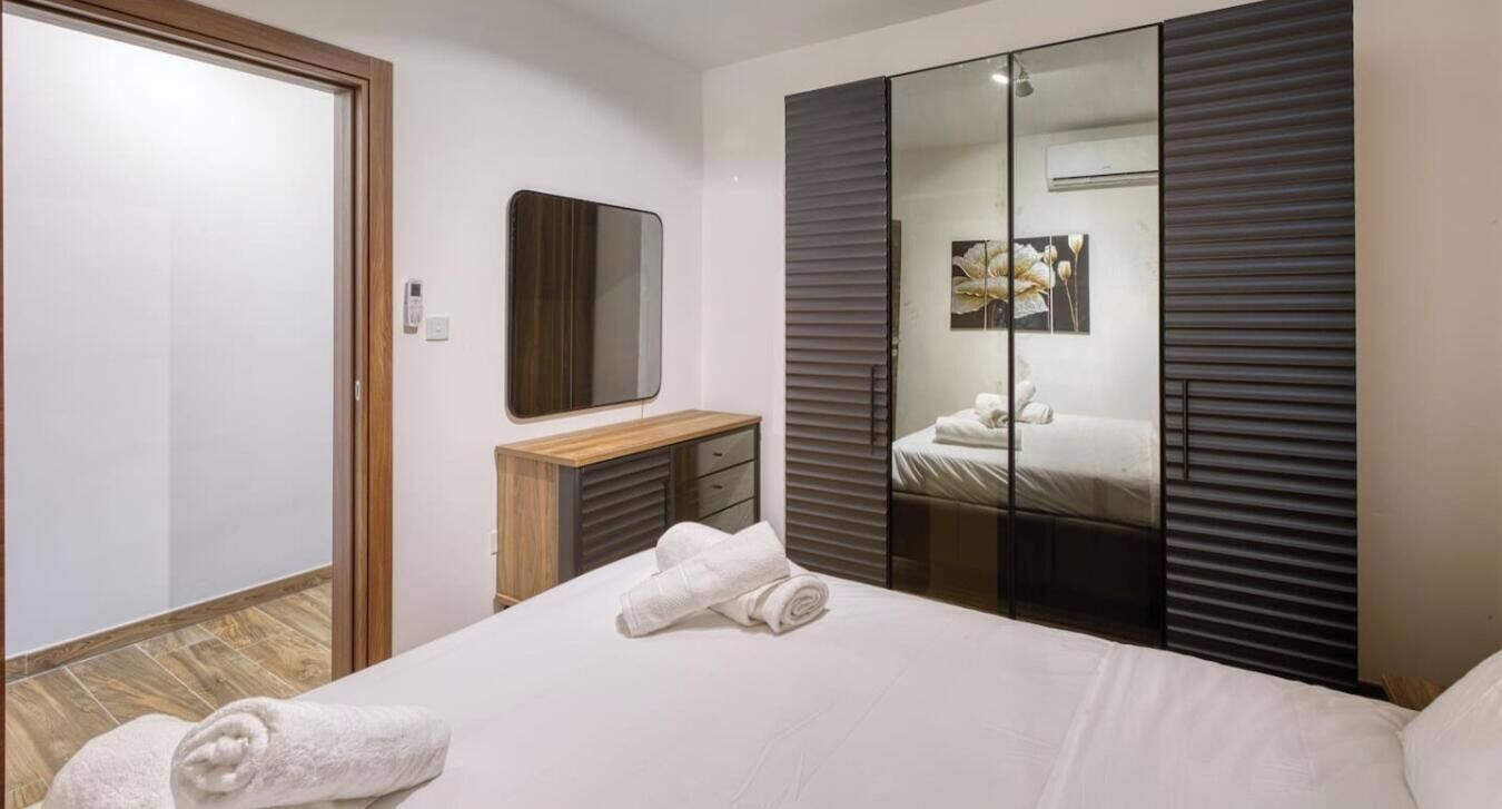 A hotel room with a bed, a wooden bedside table, a mirror, a wardrobe with a mirror, and a towel on the bed.