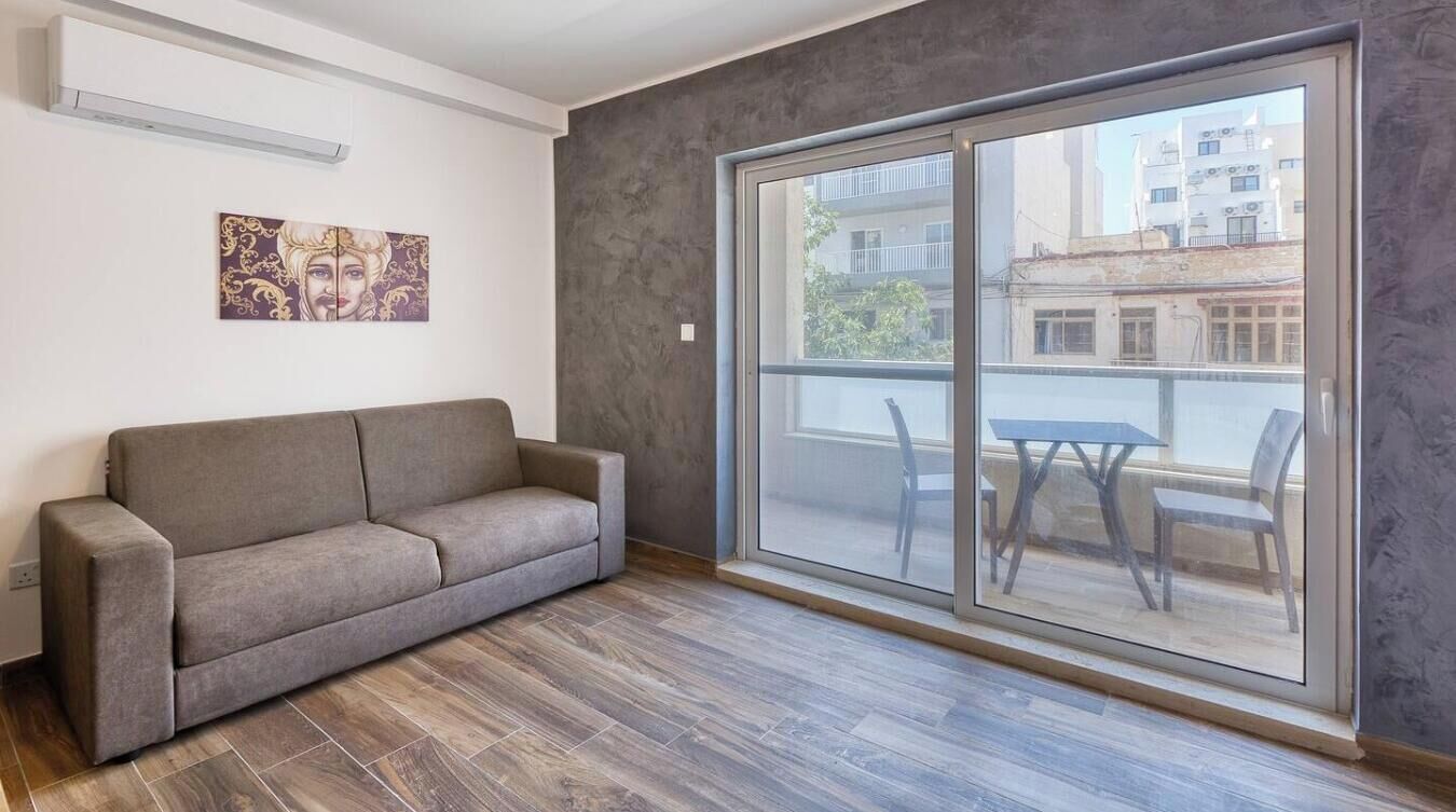 Una sala de estar moderna con un sofá gris, un amplio ventanal corredero que da a un balcón exterior y un aire acondicionado instalado en la pared.
