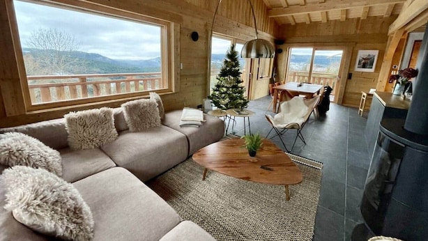 A cozy cabin interior with a large window, a sofa with fluffy pillows, a small Christmas tree, a dining table with chairs, and a fireplace. - Ma Biche: a beautiful chalet with a lake view and a Nordic hot tub! (Gérardmer)