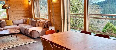 A wooden interior with a large dining table, a sofa, and large windows.