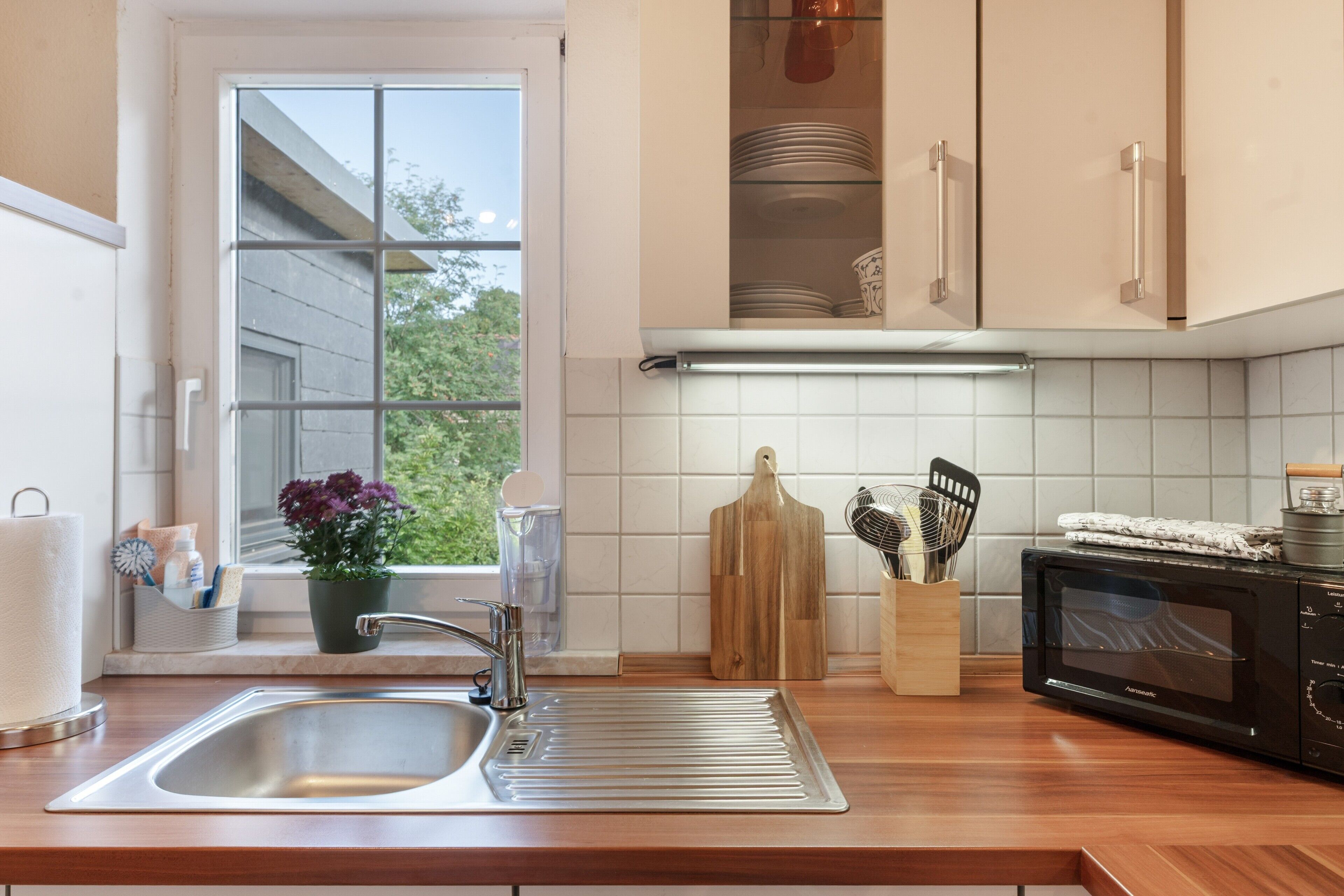 Una cocina con un lavamanos de acero inoxidable, encimera de madera, un microondas y una ventana con vista a la vegetación.