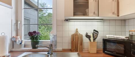 A kitchen with a stainless steel sink, wooden countertop, a microwave, and a window with a view of greenery.
