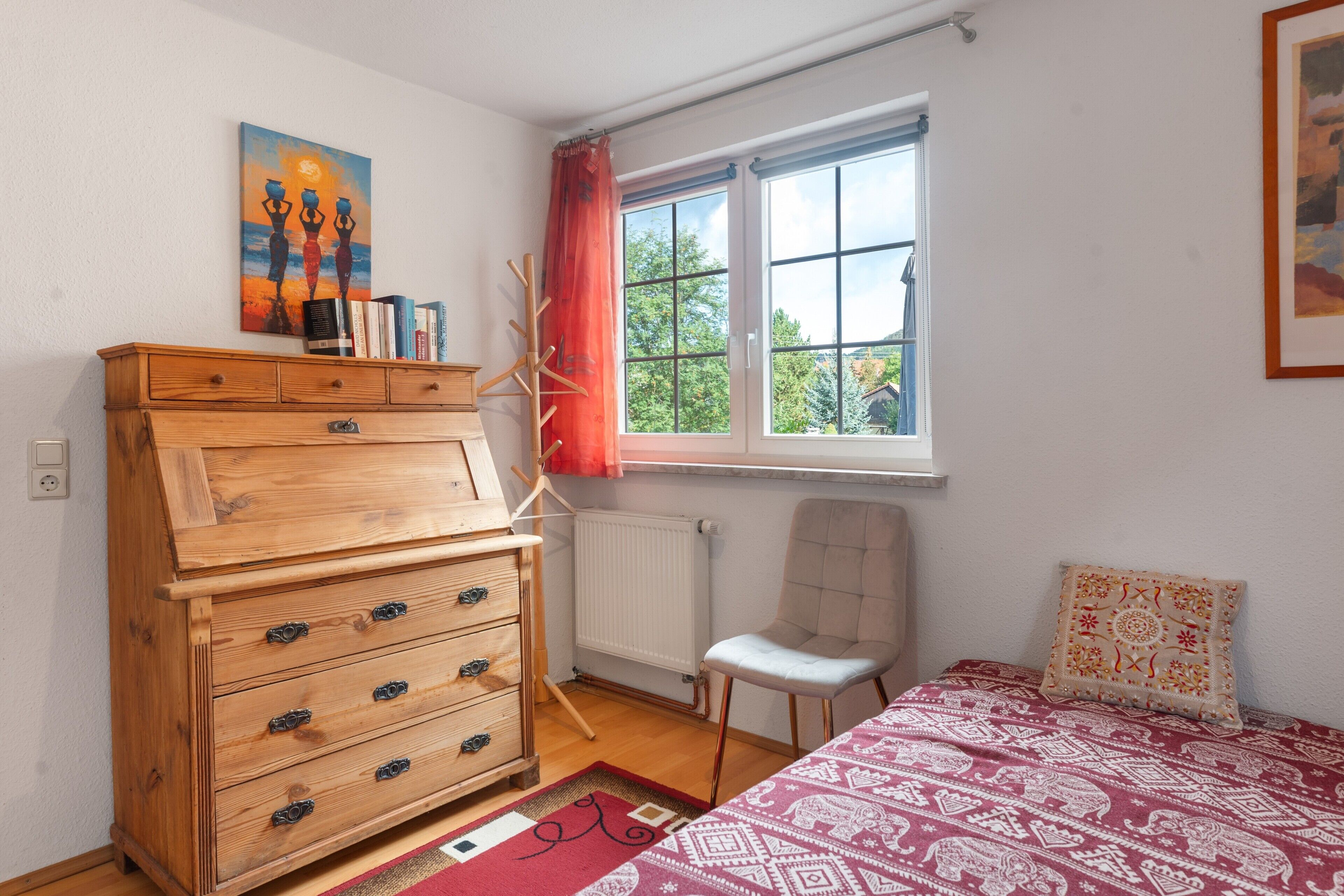 Un dormitorio con un aparador de madera, una silla, una cama con ropa de cama estampada, una ventana con vistas a árboles y un cuadro en la pared.