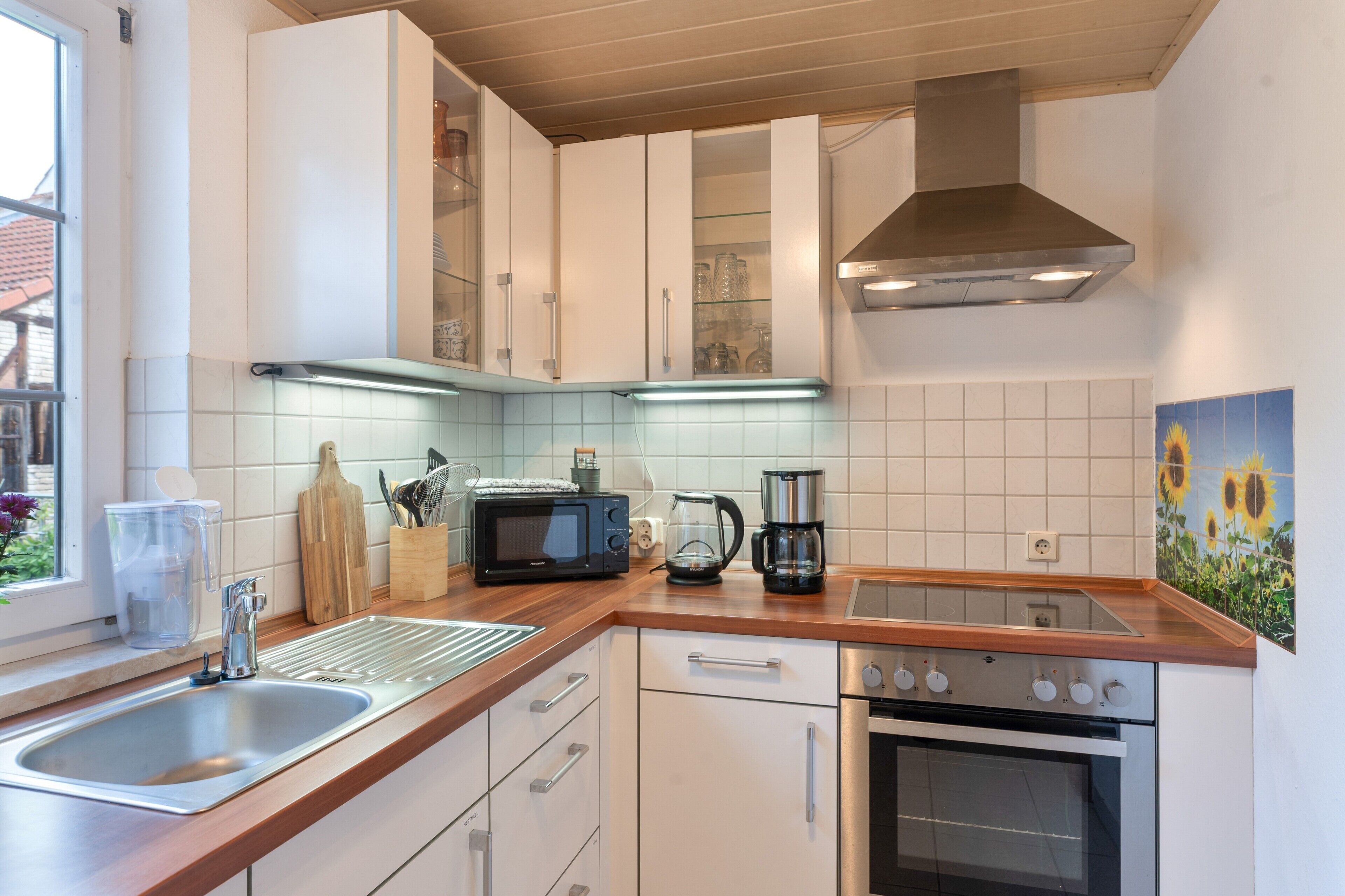Una cocina moderna con armarios blancos, un lavamanos de acero inoxidable, un microondas, una cafetera y un horno.