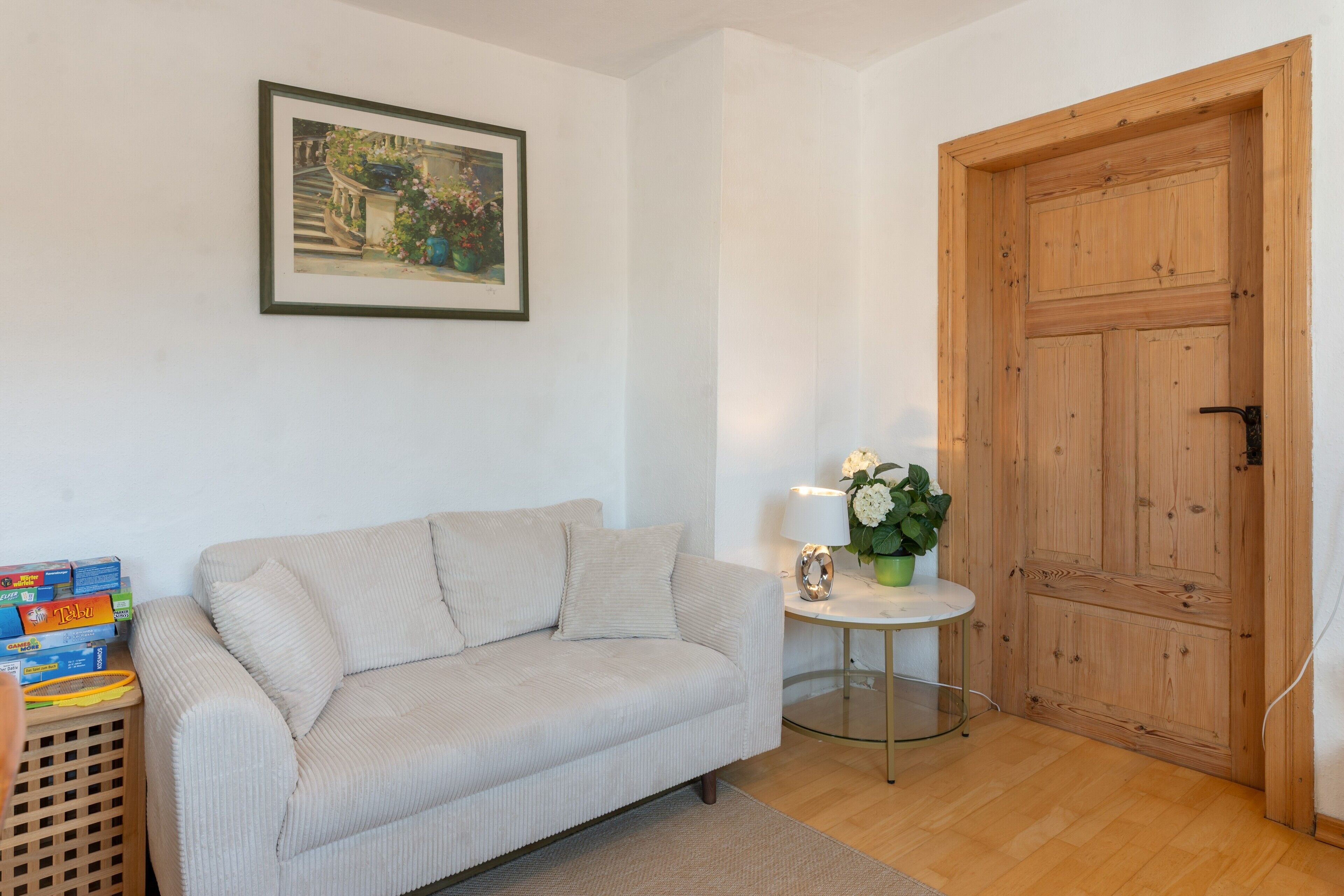 Una habitación con un sofá color beige, una mesita redonda con una lámpara, una puerta de madera y un cuadro enmarcado en la pared.