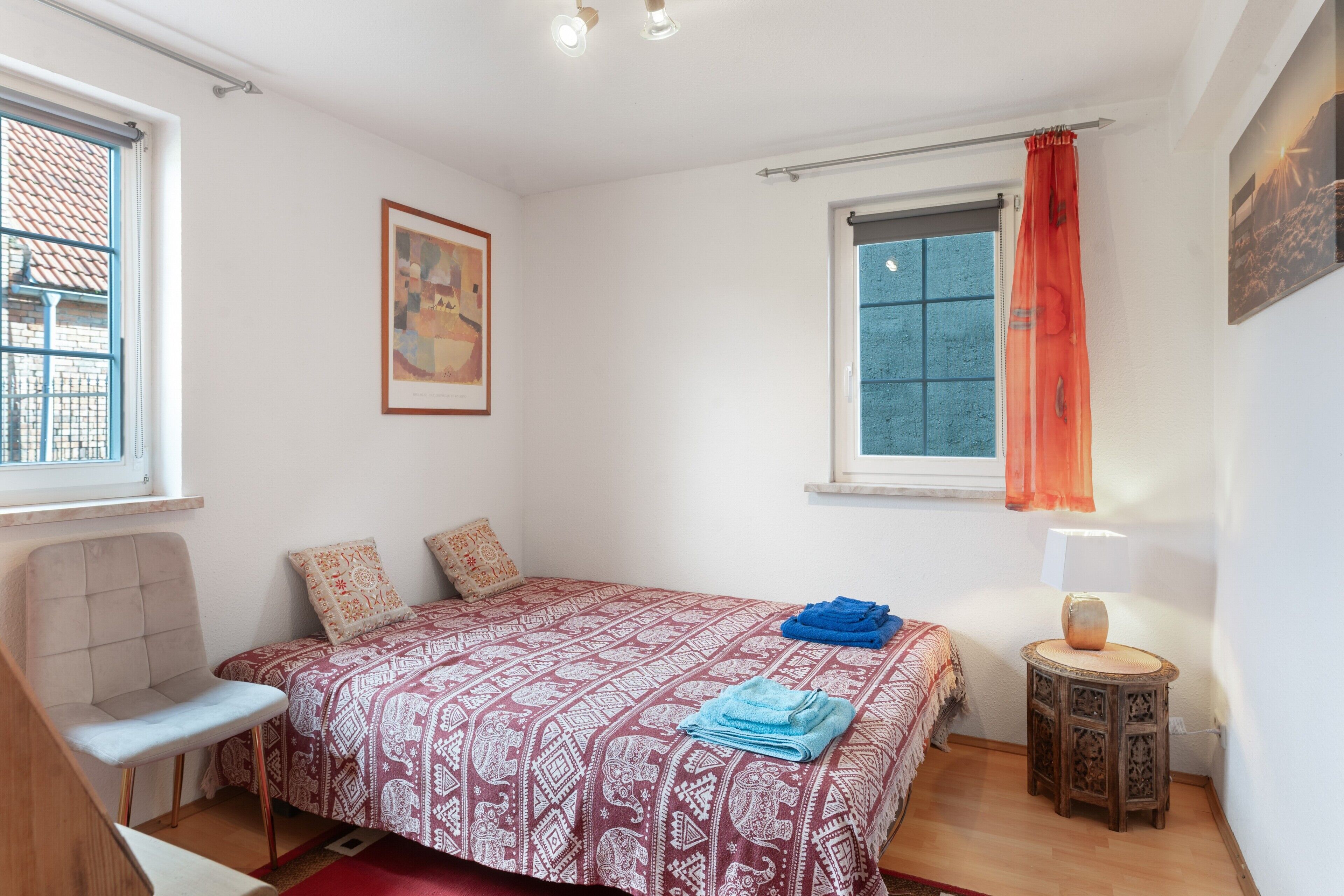 Ein Schlafzimmer mit einem Bett, einem Sessel, einem Fenster mit Vorhang, einem Nachttisch und einer Wandlampe.