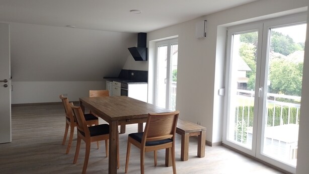 A spacious dining area with a wooden table and chairs, a kitchen in the background, and large windows with a view of greenery.