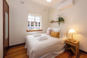 A neatly made bed with folded towels, a bedside lamp, and a window with a view of buildings outside. - Charming Art Deco Unit in East Melbourne (East Melbourne)