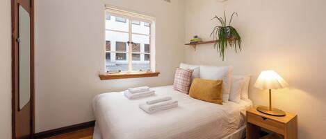 A neatly made bed with folded towels, a bedside lamp, and a window with a view of buildings outside.