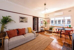 Ein Wohnzimmer mit Sofa, Esstisch mit Stühlen, großem Fenster und einem Blick sowie einer Zimmerpflanze.