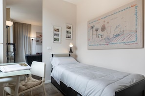Ein Einzelbett mit weißer Decke, ein Schreibtisch mit Stuhl, eine Lampe und gerahmte Bilder an der Wand.