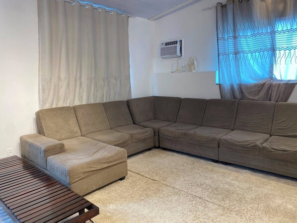 A living room with a sectional sofa, a small ottoman, a wooden coffee table, a wall-mounted air conditioner, and a window with sheer curtains.