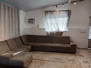 A living room with a sectional sofa, a window with sheer curtains, an air conditioning unit, and a patterned carpet.