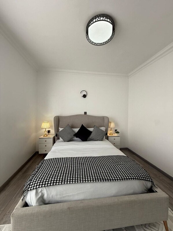 A neatly arranged bedroom with a bed, bedside tables, lamps, a round mirror, and a wall-mounted light fixture.
