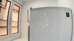 Ett kompakt kök med en inbyggd tvättmaskin, ett kylskåp och ett fönster med persienner.