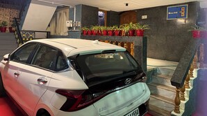 A white car parked indoors with a staircase, potted plants, and a blue sign on the wall. - Eden Gardens Home Stay (Coimbatore)