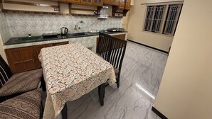 A kitchen with wooden cabinets, a marble countertop, a table with a lace tablecloth, and a window with bars. - Eden Gardens Home Stay (Coimbatore)