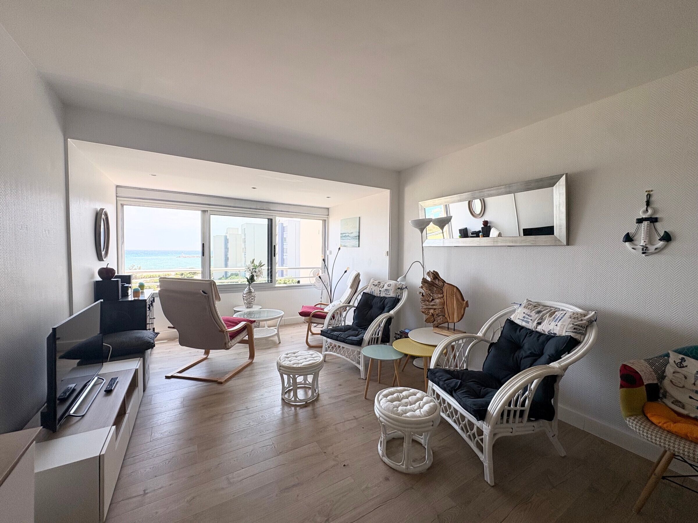 A modern living room with a view of the sea, furnished with white wicker chairs, a sofa, and a small table.