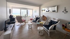 Un salon moderne avec vue sur la mer, aménagé avec des fauteuils en osier blanc, un canapé et une petite table.