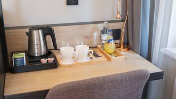Une chambre d’hôtel avec un bureau en bois, une machine à café, deux tasses blanches, un plateau avec une bouteille et des verres, et une lampe.