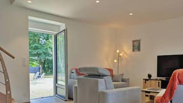 Un salon moderne avec un canapé, un téléviseur et une lampe sur pied. Une porte-fenêtre coulissante donne accès à un espace extérieur agrémenté d’un banc.