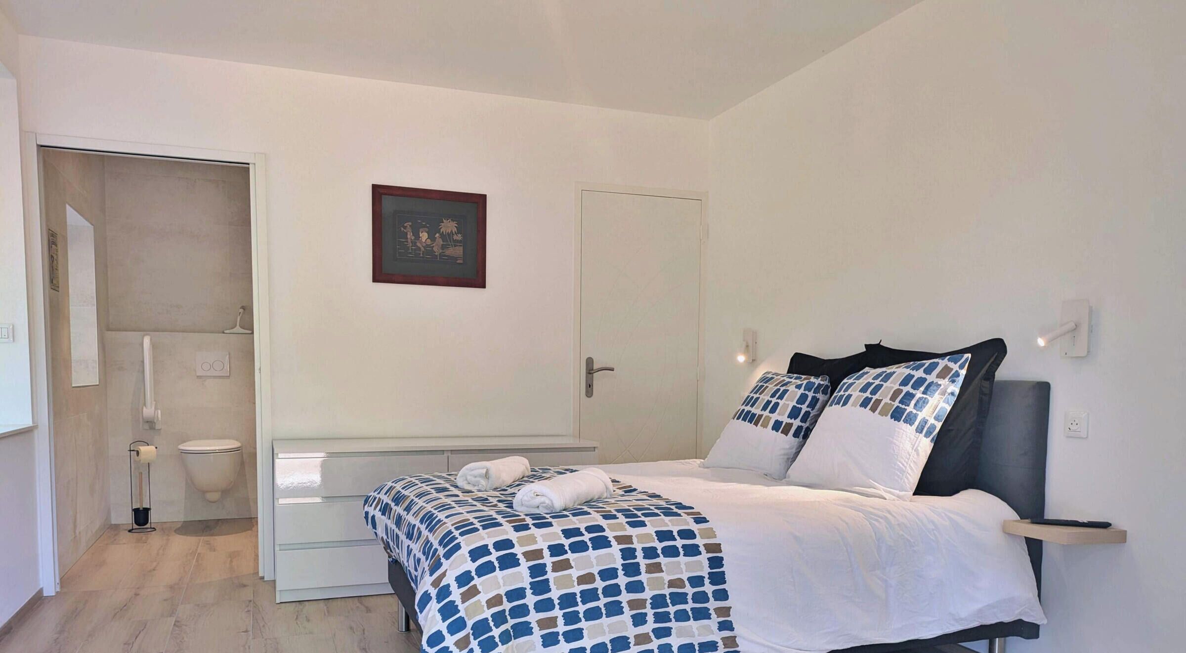 A bedroom with a bed, a bedside table, a nightstand, a door, and a framed picture on the wall.