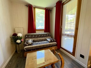 Ein Eckzimmer mit einer Couch, einem kleinen Tisch, einem Fenster mit roten Vorhängen und Blick ins Grüne.