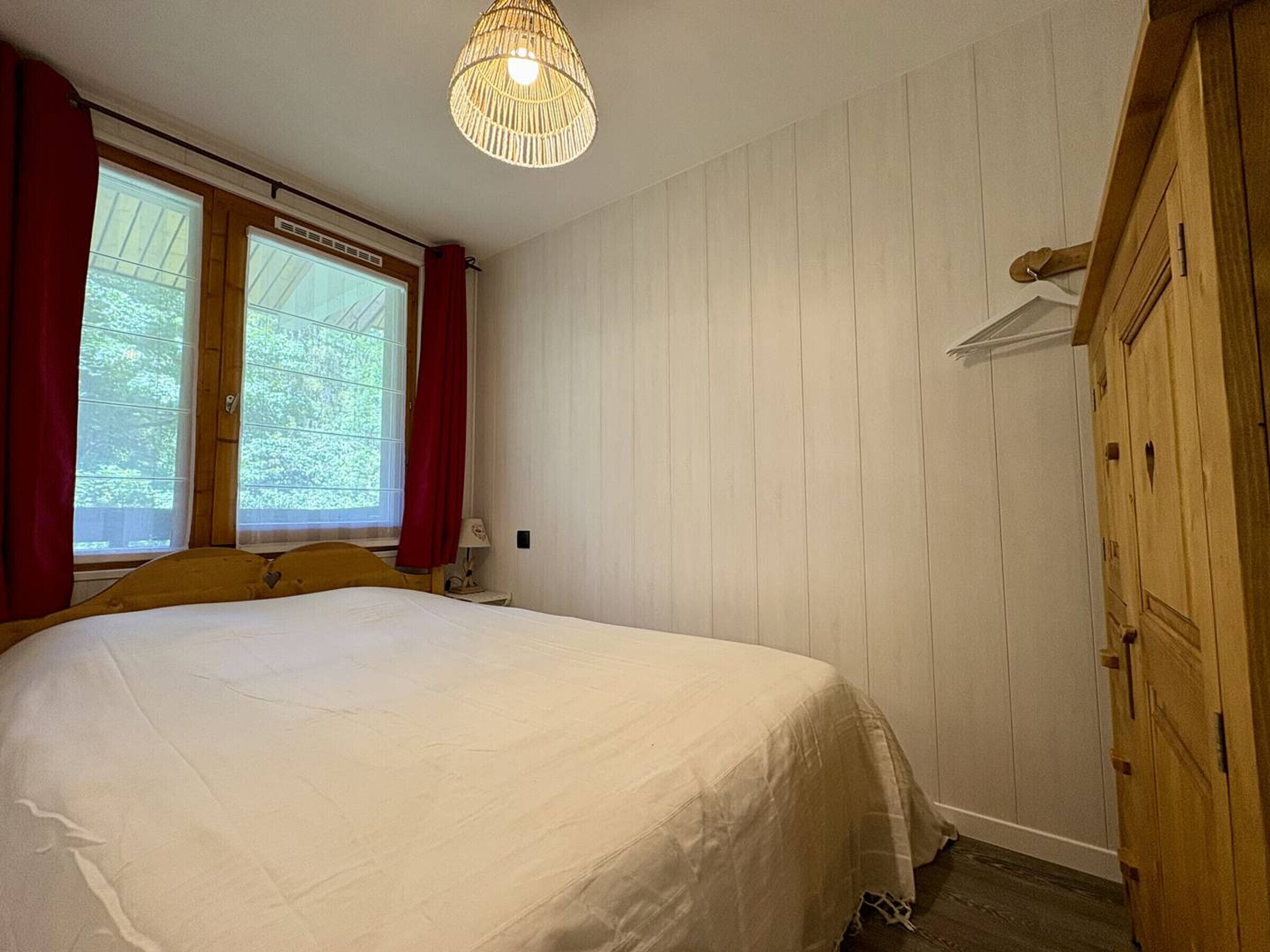 A bedroom with a bed, a wooden wardrobe, a window with curtains, and a hanging light fixture.